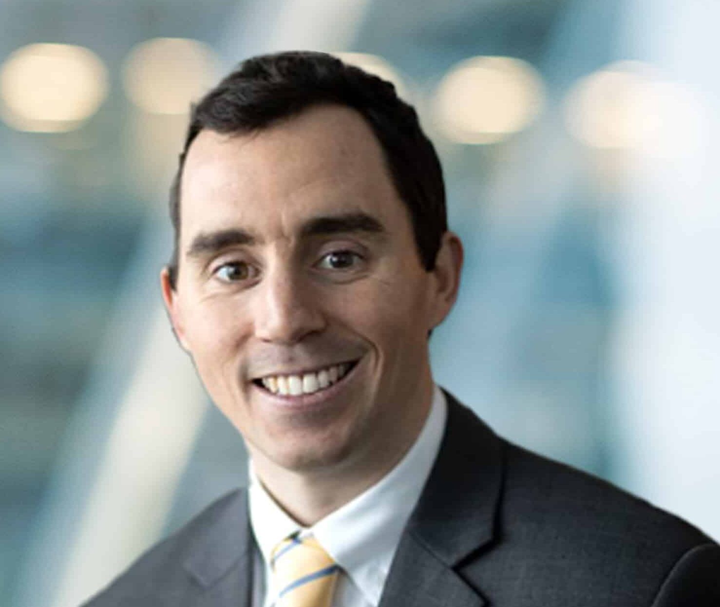 A man in a dark suit, white shirt, and striped tie smiles at the camera with a blurred, modern office background.