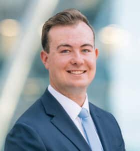 A young man in a navy suit, white shirt, and light blue tie smiles at the camera. The background is blurred with soft lights and a modern, glass building.