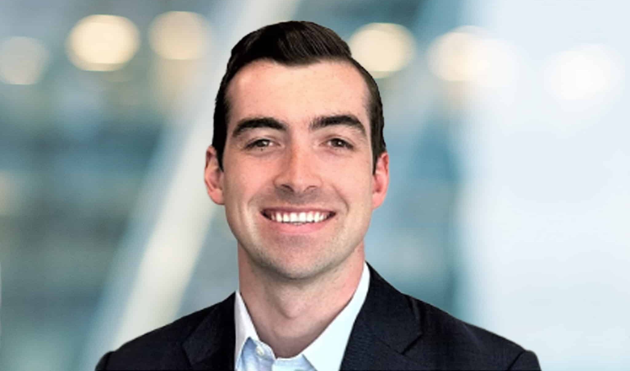 A man with dark hair wearing a dark blazer and white shirt smiles at the camera. The background is blurred with soft lights and shades of blue and gray.