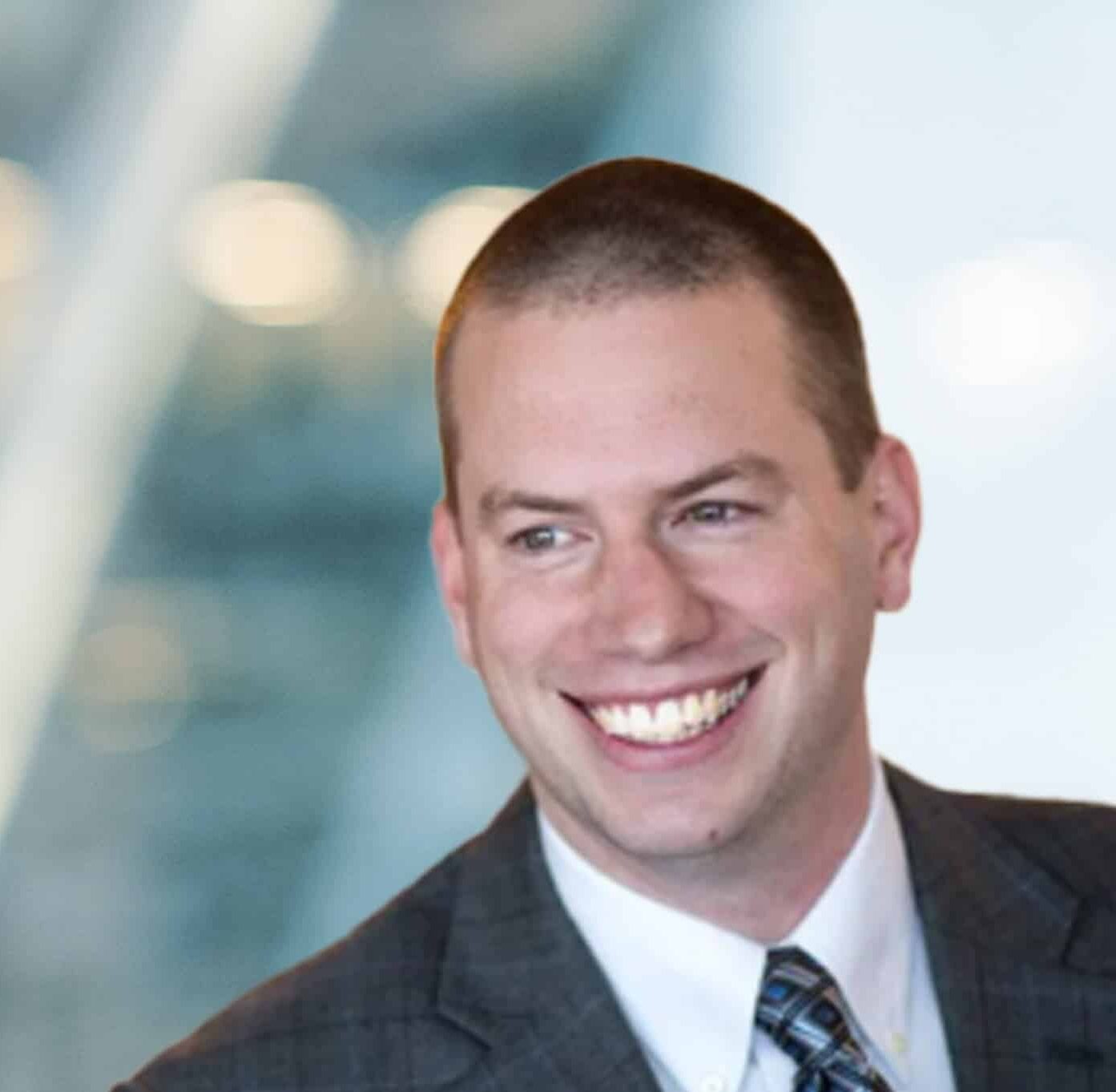 A man in a gray plaid suit and striped tie smiles broadly, looking to the side, with a blurred modern office background behind him.