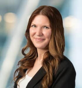 A woman with long, wavy brown hair wearing a black blazer and white top smiles at the camera, with a blurred, modern office background.