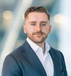 A man with short brown hair and a trimmed beard, wearing a dark suit jacket and white shirt, stands in front of a blurred, modern office background with soft lighting.