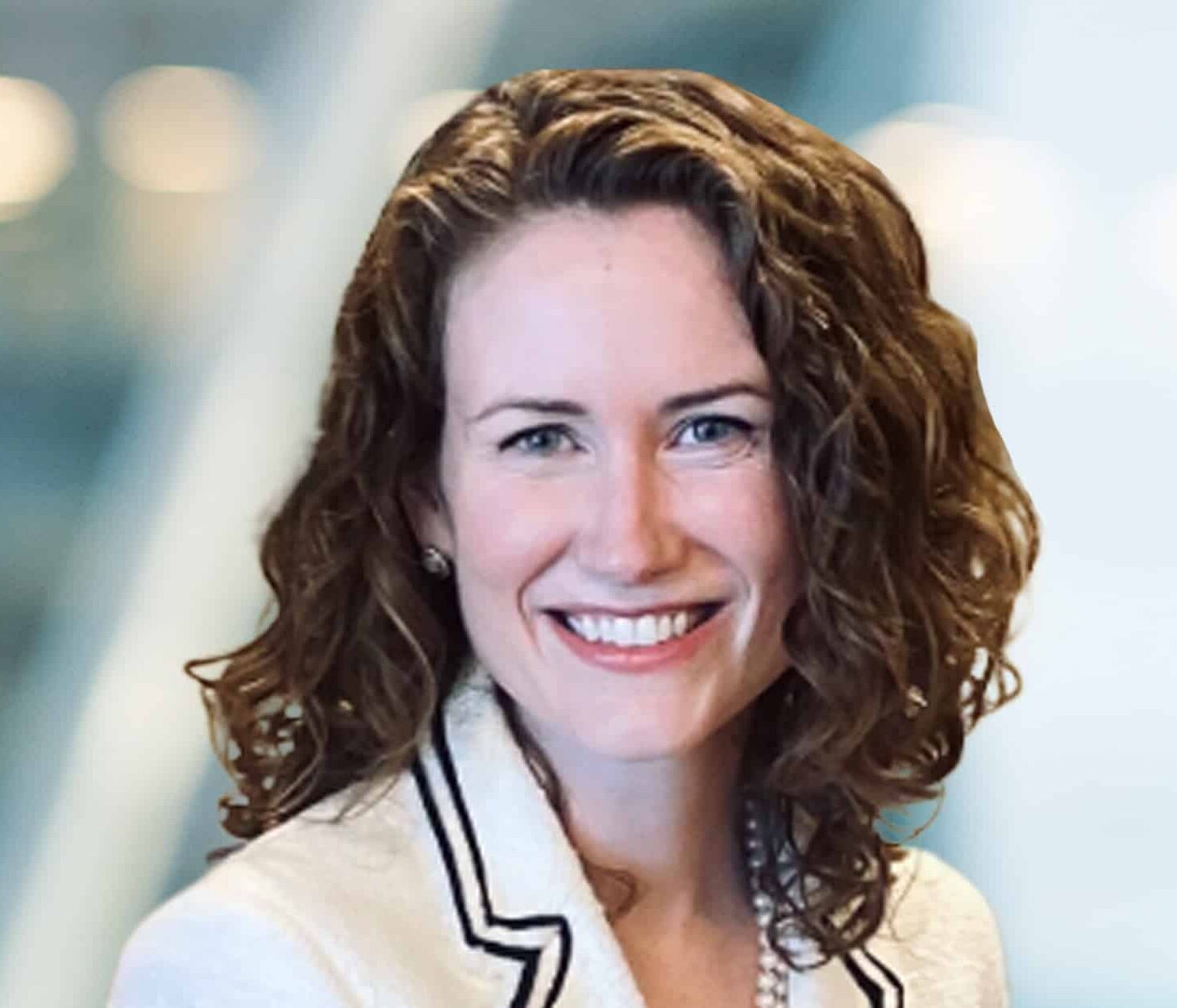 A woman with curly brown hair, wearing a white blazer and pearl necklace, smiles at the camera against a soft, blurred background.