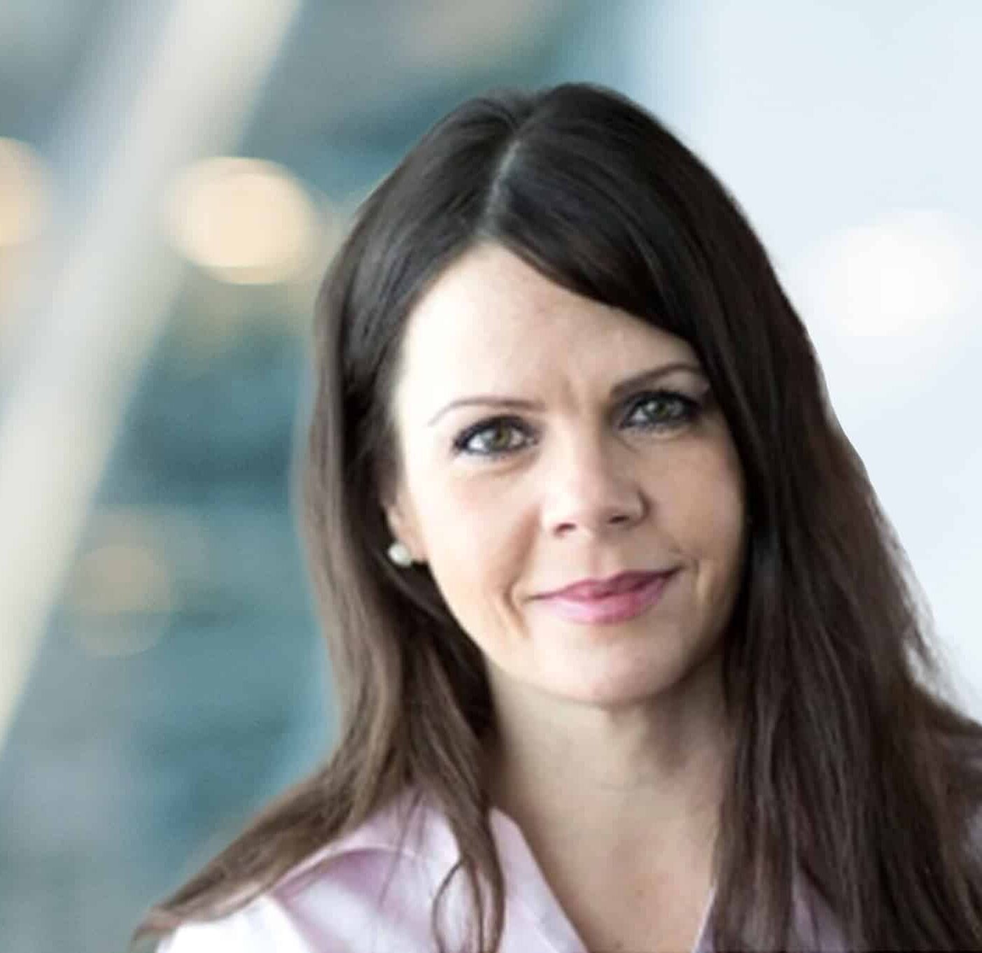 A woman with long brown hair and a light pink shirt smiles softly, standing in front of a blurred modern office background with soft lighting.