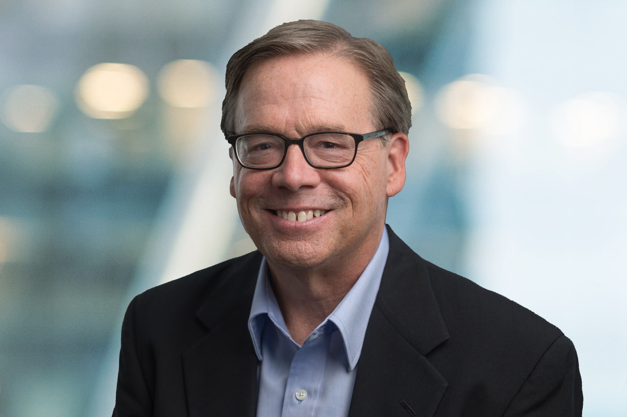 A middle-aged man with light brown hair and glasses, wearing a dark blazer and light blue shirt, smiles at the camera against a blurred background with soft lights.