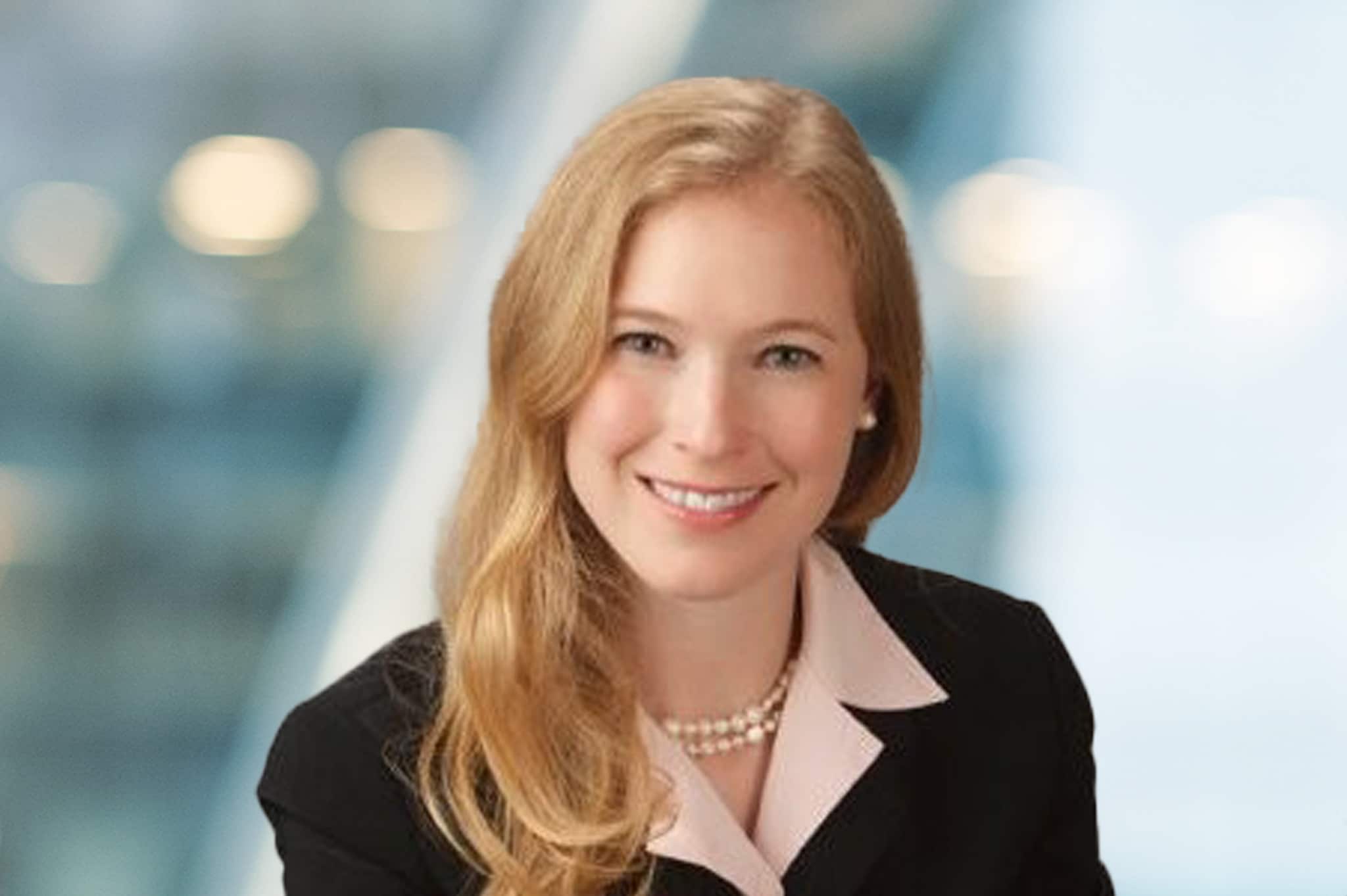 A woman with long blonde hair wearing a black blazer, light pink blouse, and pearl necklace, smiling in front of a blurred office background.