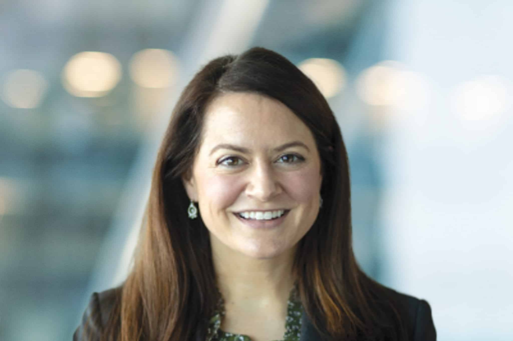 A woman with long brown hair, wearing a dark blazer and green necklace, smiles at the camera. The background is blurred with soft lights and blue tones.