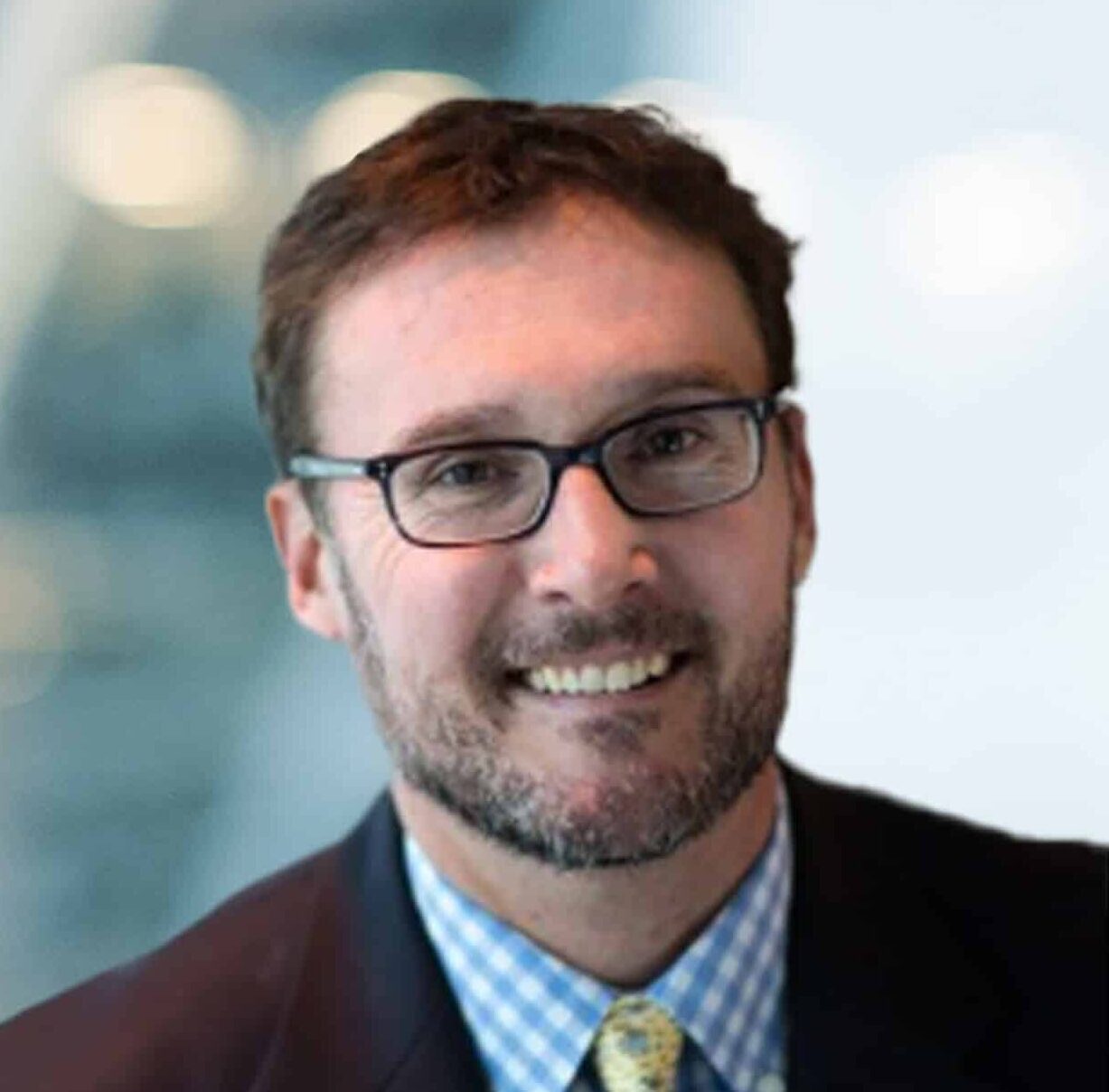 A man with brown hair, a beard, and glasses, wearing a suit jacket, blue checked shirt, and patterned tie, smiles in front of a blurred modern office background with lights.
