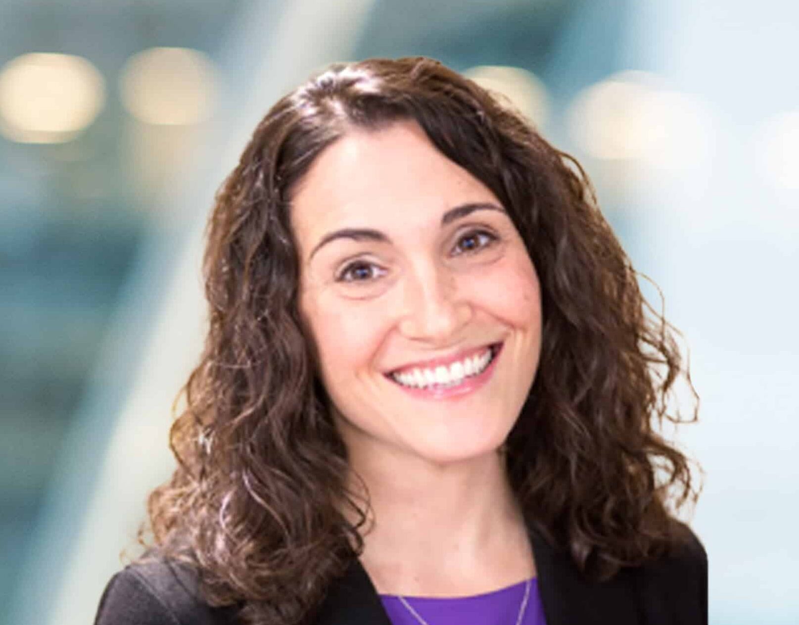 A woman with curly brown hair and a bright smile stands in front of a blurred, modern indoor background. She is wearing a black blazer over a purple top.