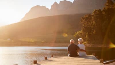 An elderly couple sits together on a wooden dock overlooking a calm lake, surrounded by trees and mountains, with warm sunlight illuminating the peaceful landscape.