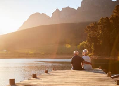 An elderly couple sits together on a wooden dock overlooking a calm lake, surrounded by trees and mountains, with warm sunlight illuminating the peaceful landscape.