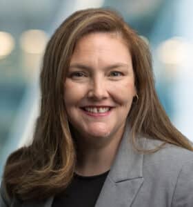 A woman with long brown hair wearing a gray blazer and black top smiles at the camera. The background is softly blurred with blue and white tones and circular lights.