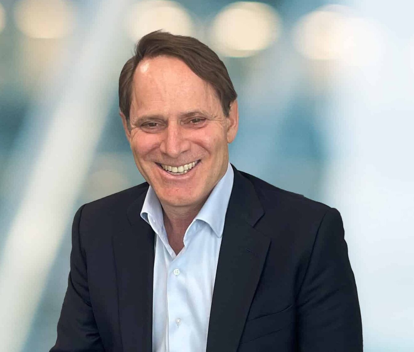 A man with short brown hair wearing a dark suit jacket and light blue shirt smiles in front of a blurred, modern office background with blue and white tones.